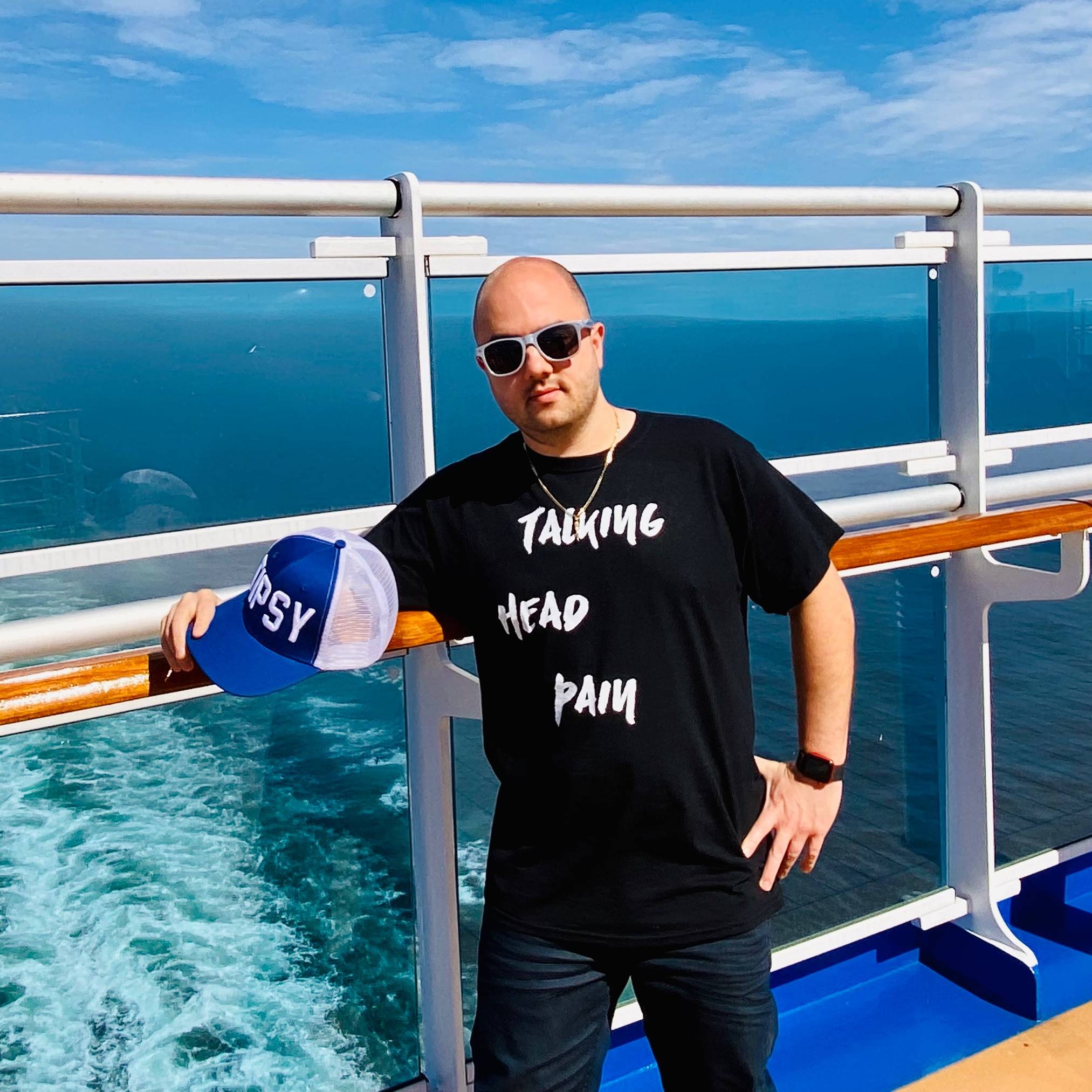 A Photo Of Joe Coe, MPA, Director Of Education And Digital Strategy At GHLF, Wearing A Black T-shirt That Has The Words "Talking Head Pain" Written In White Font. Joe Is Wearing A Pair Of White Sunglasses Which Were Designed By Michael Kuluva, Fashion Designer And Creator Of The Fashion Sportswear Brand Tumbler & Tipsy. The Sunglasses Were Designed To Raise Awareness Around Migraine.